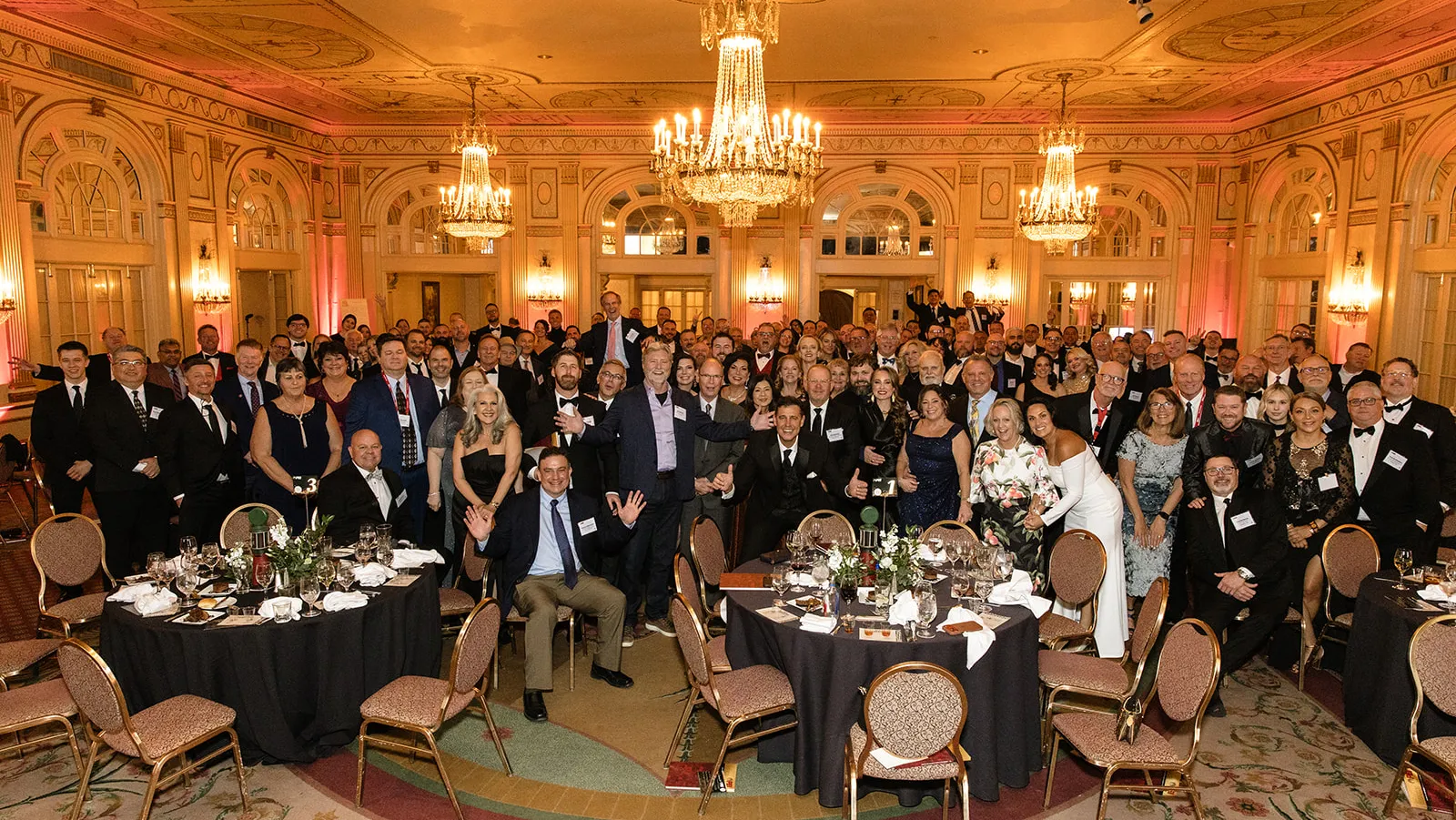 A large group of formally dressed people gathers in an elegant ballroom with chandeliers, round tables set with glasses and plates, and ornate walls, posing for a group photo at the PEI 75th anniversary gala.