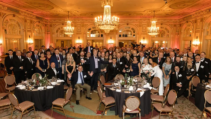 A large group of formally dressed people gathers in an elegant ballroom with chandeliers, round tables set with glasses and plates, and ornate walls, posing for a group photo at the PEI 75th anniversary gala.
