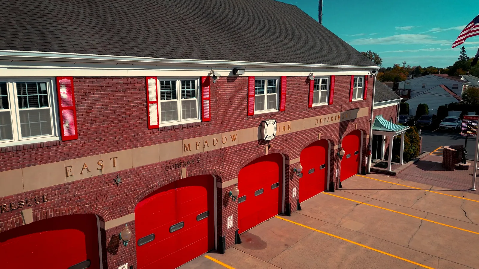 A brick fire station with bright red garage doors, labeled "East Meadow Fire Department." The building has red window shutters, an American flag on a pole, and a clear, sunny sky above.